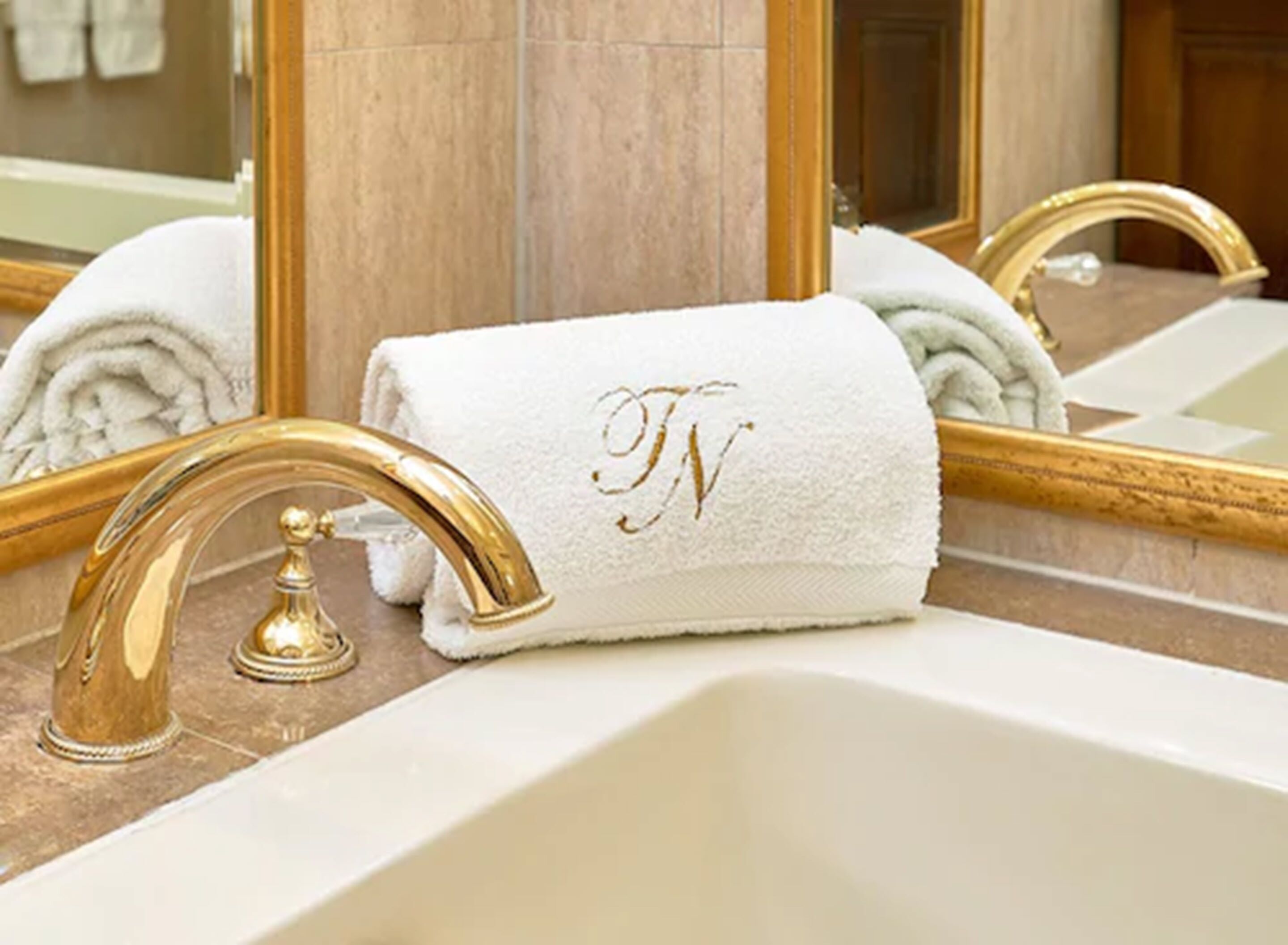 A bathroom with a gold faucet, a white sink, and a towel with a monogram.