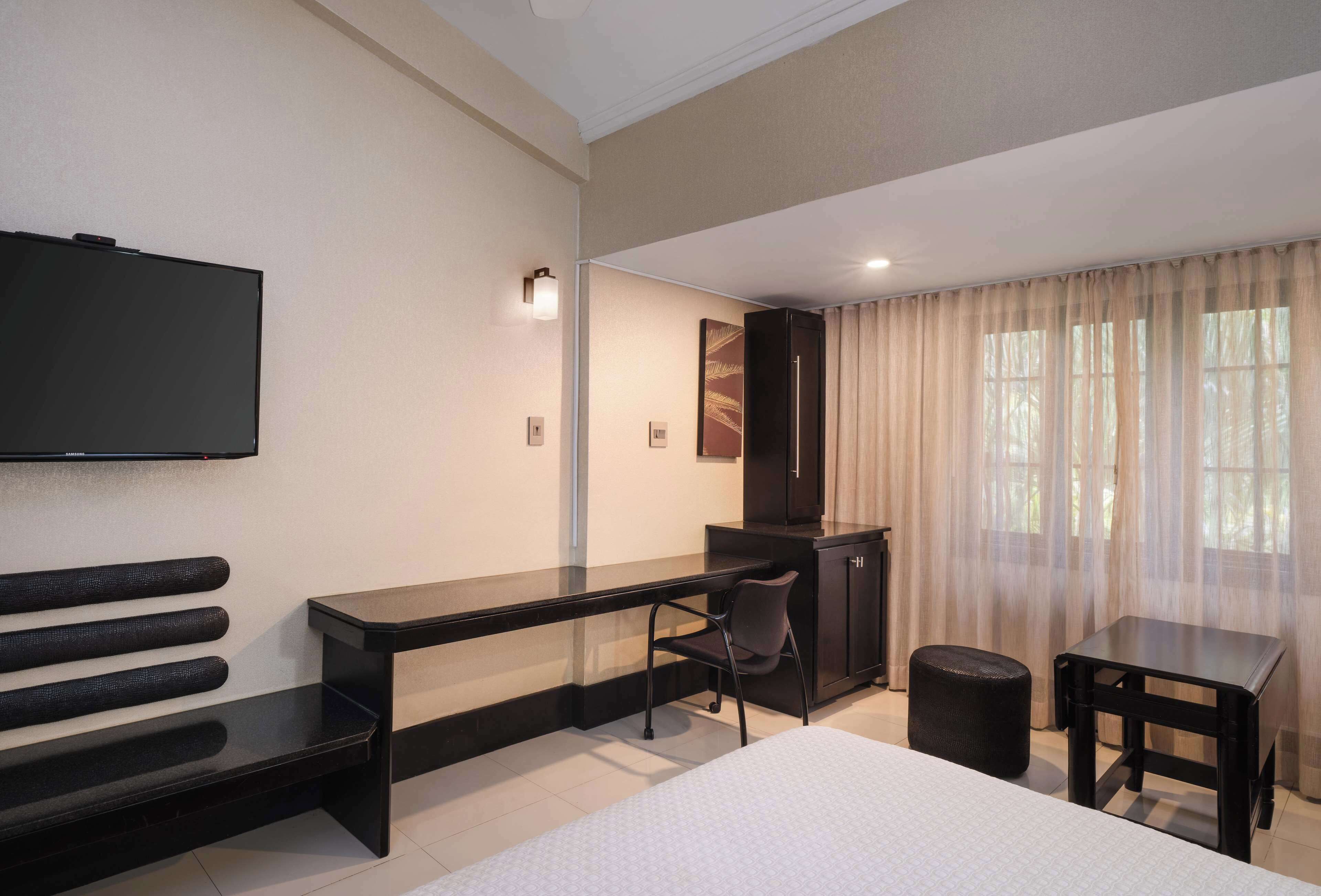 Habitación de hotel con cama, escritorio, silla, televisor, armario y ventana con cortinas.