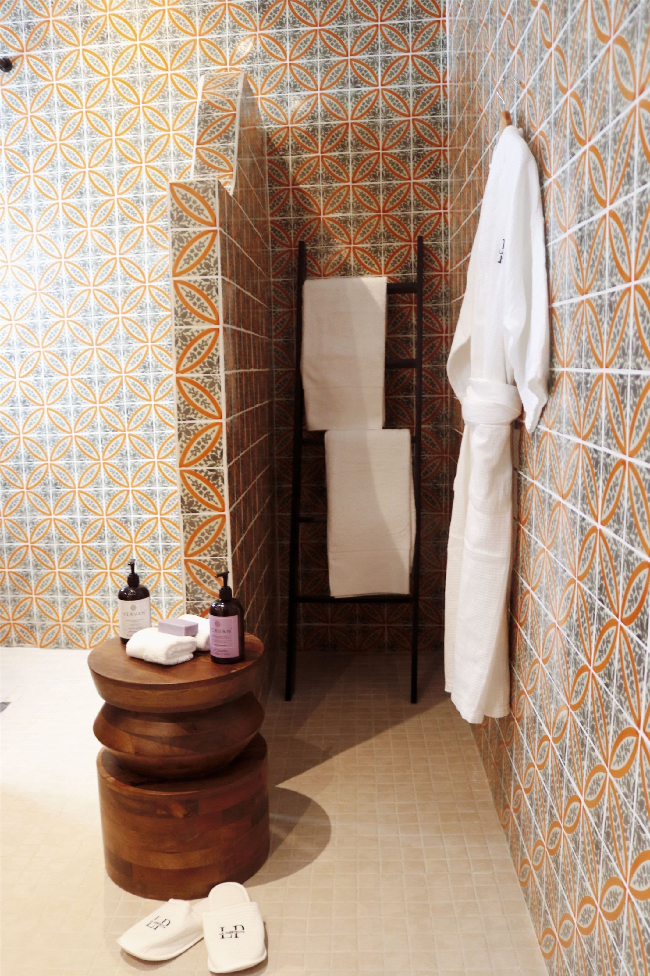 Un baño con baldosas estampadas, un taburete de madera, un perchero y una botella de loción.
