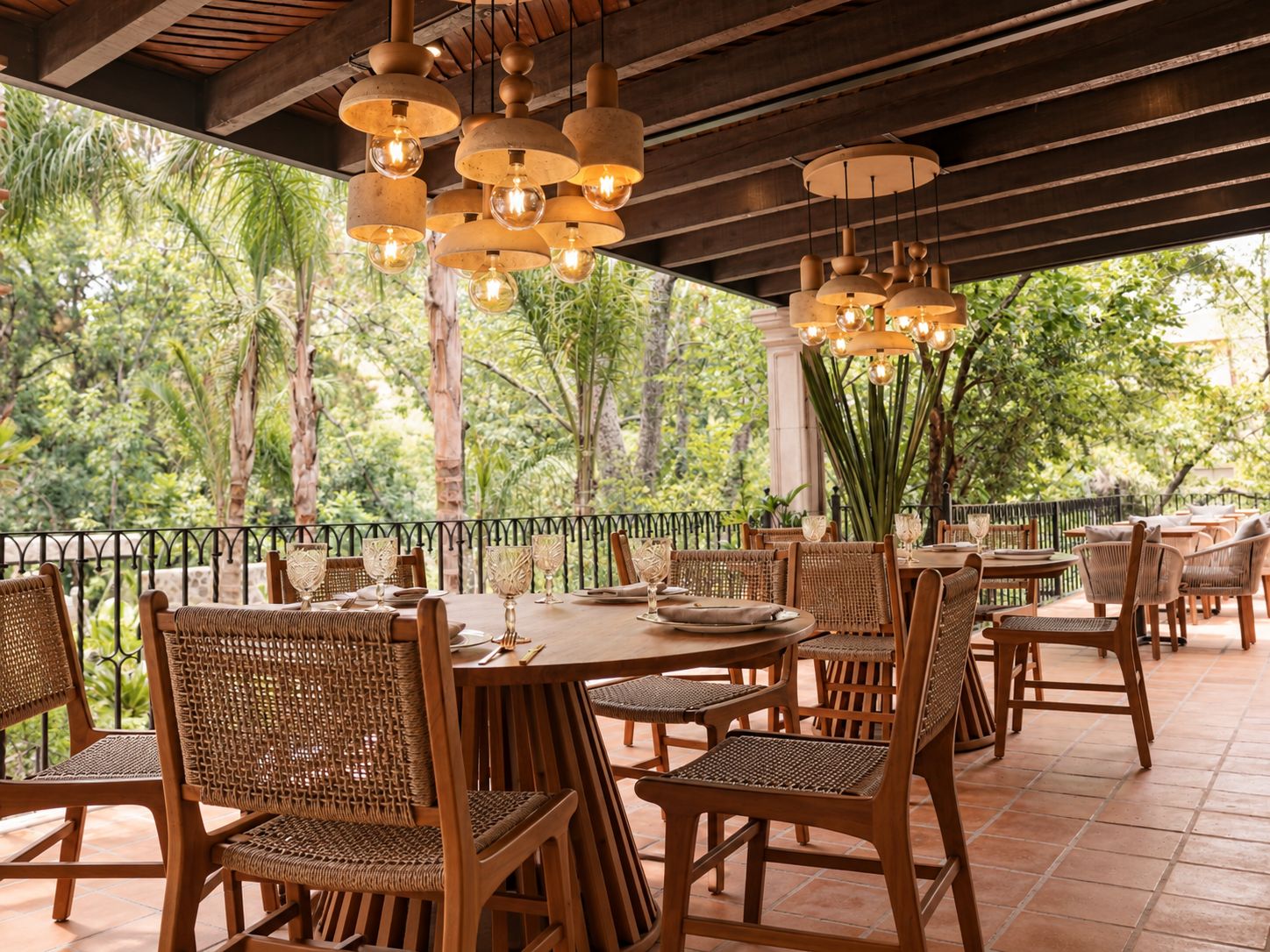 An outdoor dining area with wicker chairs and tables, surrounded by a railing, under a wooden ceiling with hanging light fixtures.