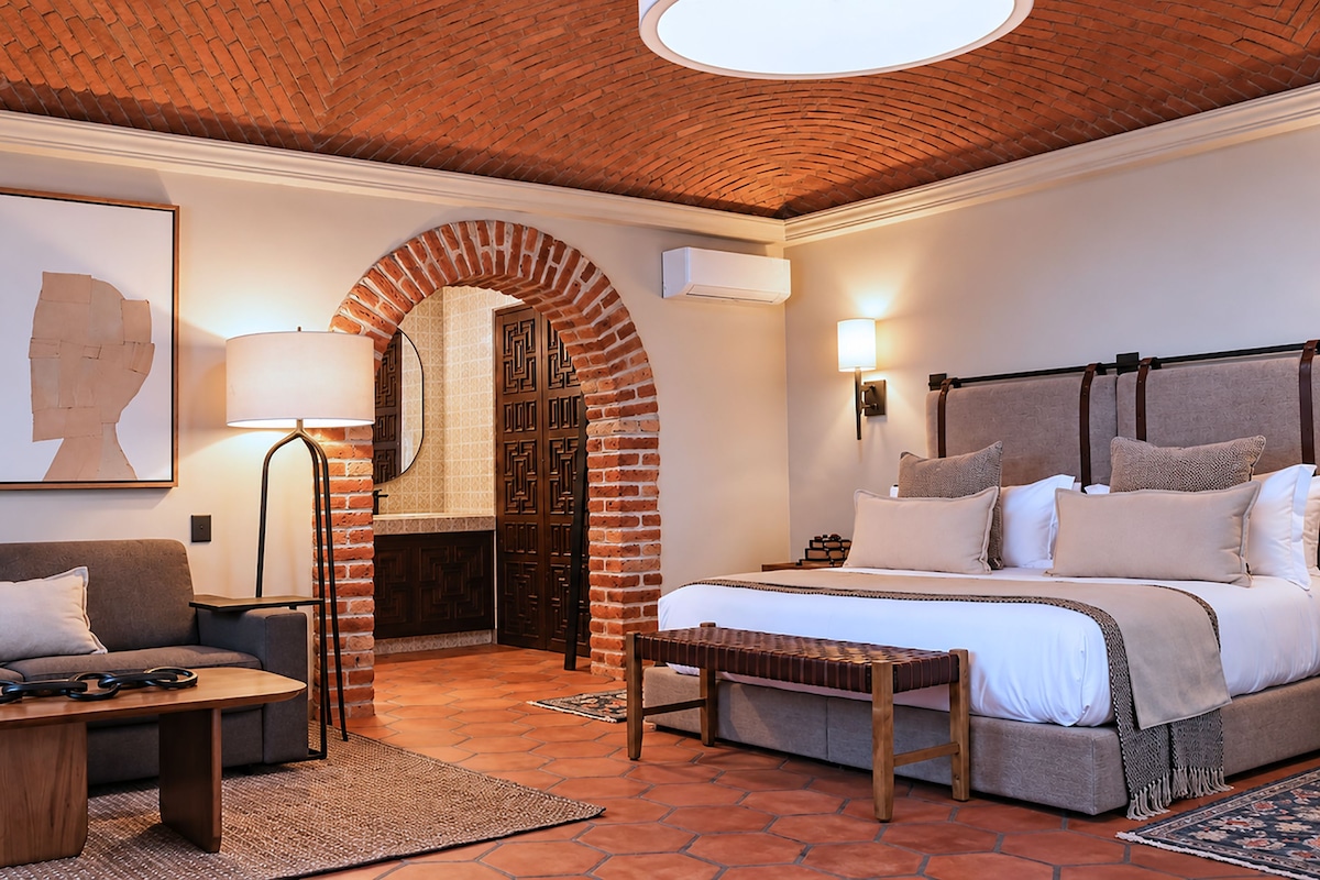 A hotel room with a brick fireplace, a bed with multiple pillows, a sofa, a coffee table, and two framed artworks.