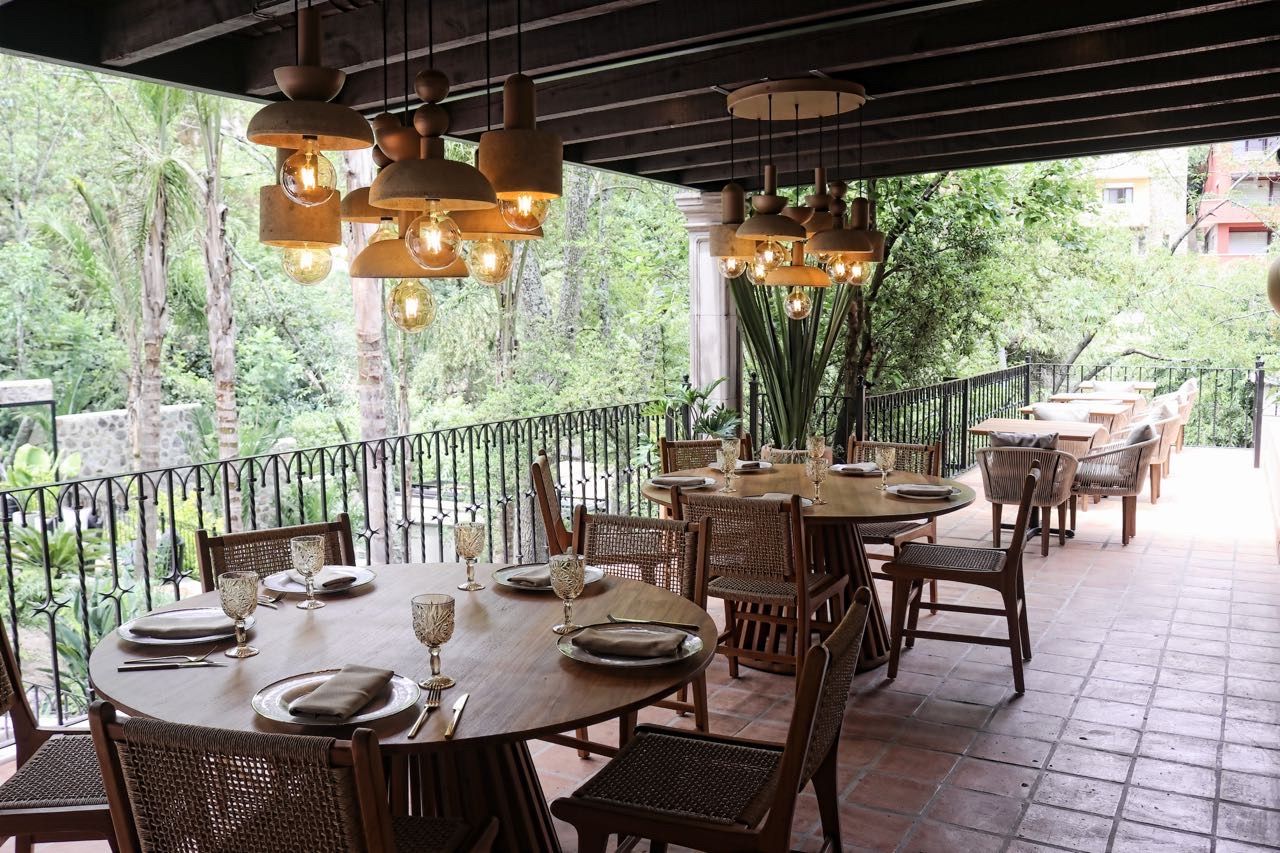 Un área de comedor al aire libre con mesas redondas y sillas de mimbre, rodeada por una baranda metálica, con vistas a un exuberante jardín.