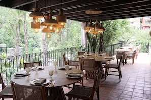 An outdoor dining area with round tables and wicker chairs, surrounded by a metal railing, overlooking a lush garden.