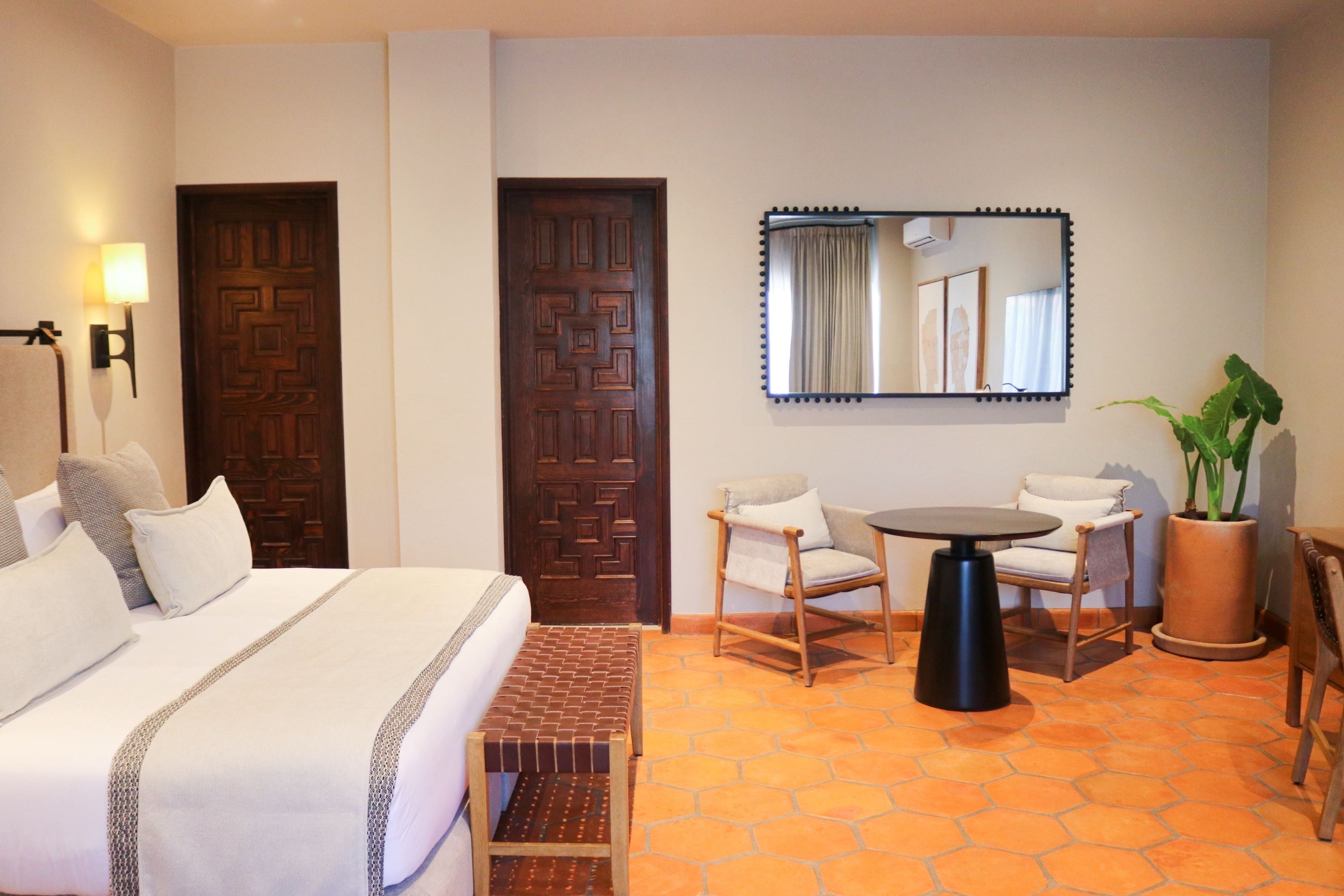 A hotel room with a bed, a small table, two chairs, a bench, a mirror, and a potted plant.