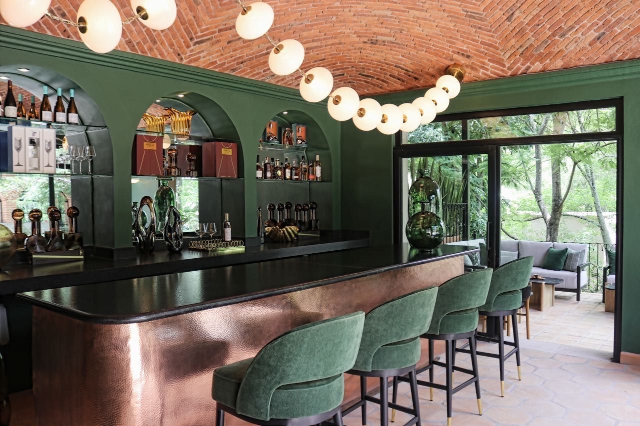 Zona de bar con mostrador de cobre, taburetes de bar tapizados en verde y techo de ladrillo con lámparas esféricas colgantes.