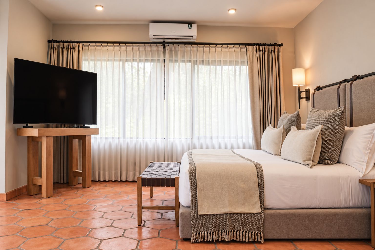 Una habitación de hotel con una cama, un televisor sobre un soporte de madera, una mesita y una ventana con cortinas.