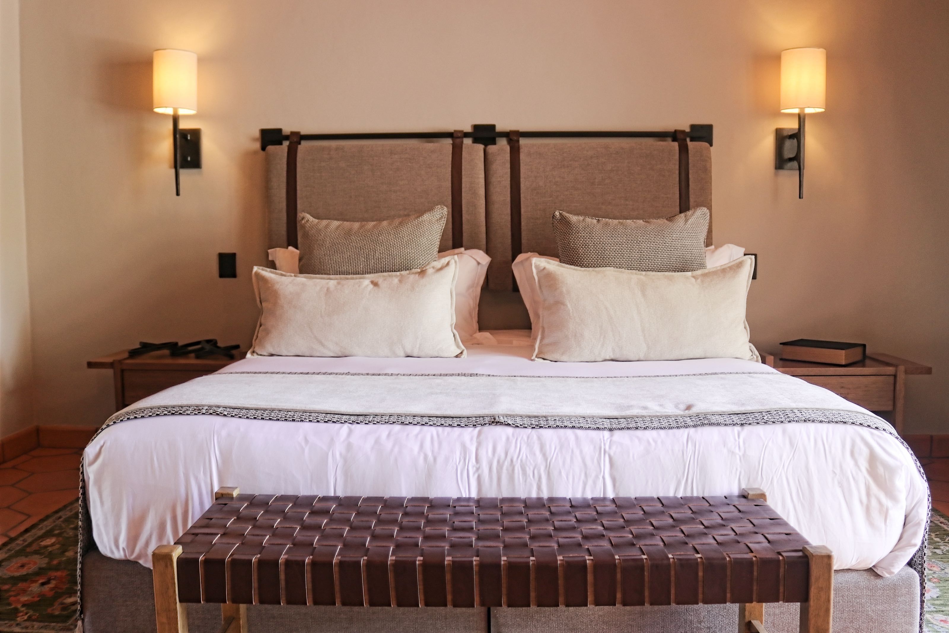 Une chambre d’hôtel avec un grand lit, deux tables de chevet, un banc et des lampes fixées au mur.