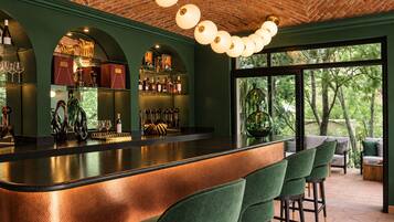 Zona de bar con barra curva, taburetes de bar tapizados en verde y techo de ladrillo con lámparas esféricas colgantes.
