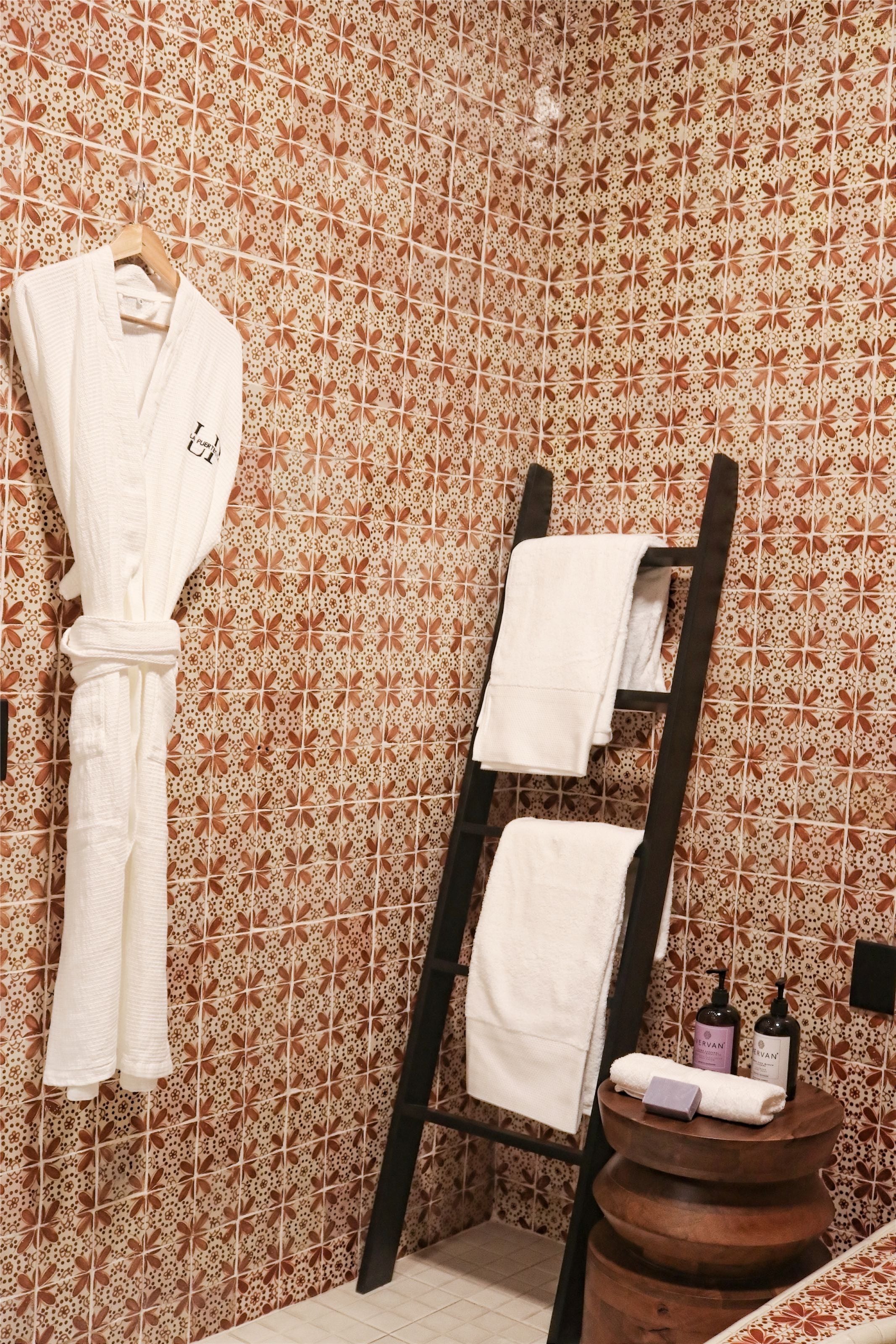 Un baño con pared de azulejos estampados, un perchero de madera y un taburete de madera con artículos de tocador.