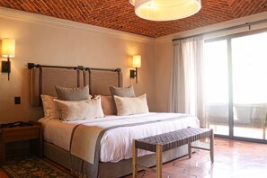 A hotel room with a brick ceiling, a large bed with multiple pillows, a nightstand, a bench, and a view of the outdoors through a large window.