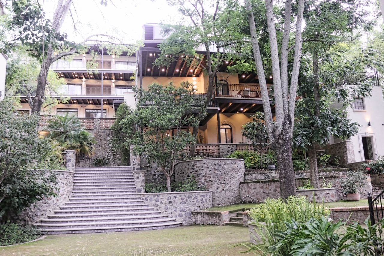 Un edificio de varios pisos con una escalera de piedra que conduce a un nivel inferior, rodeado de abundante vegetación.
