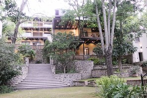 A multi-story building with a stone staircase leading to a lower level, surrounded by lush greenery.