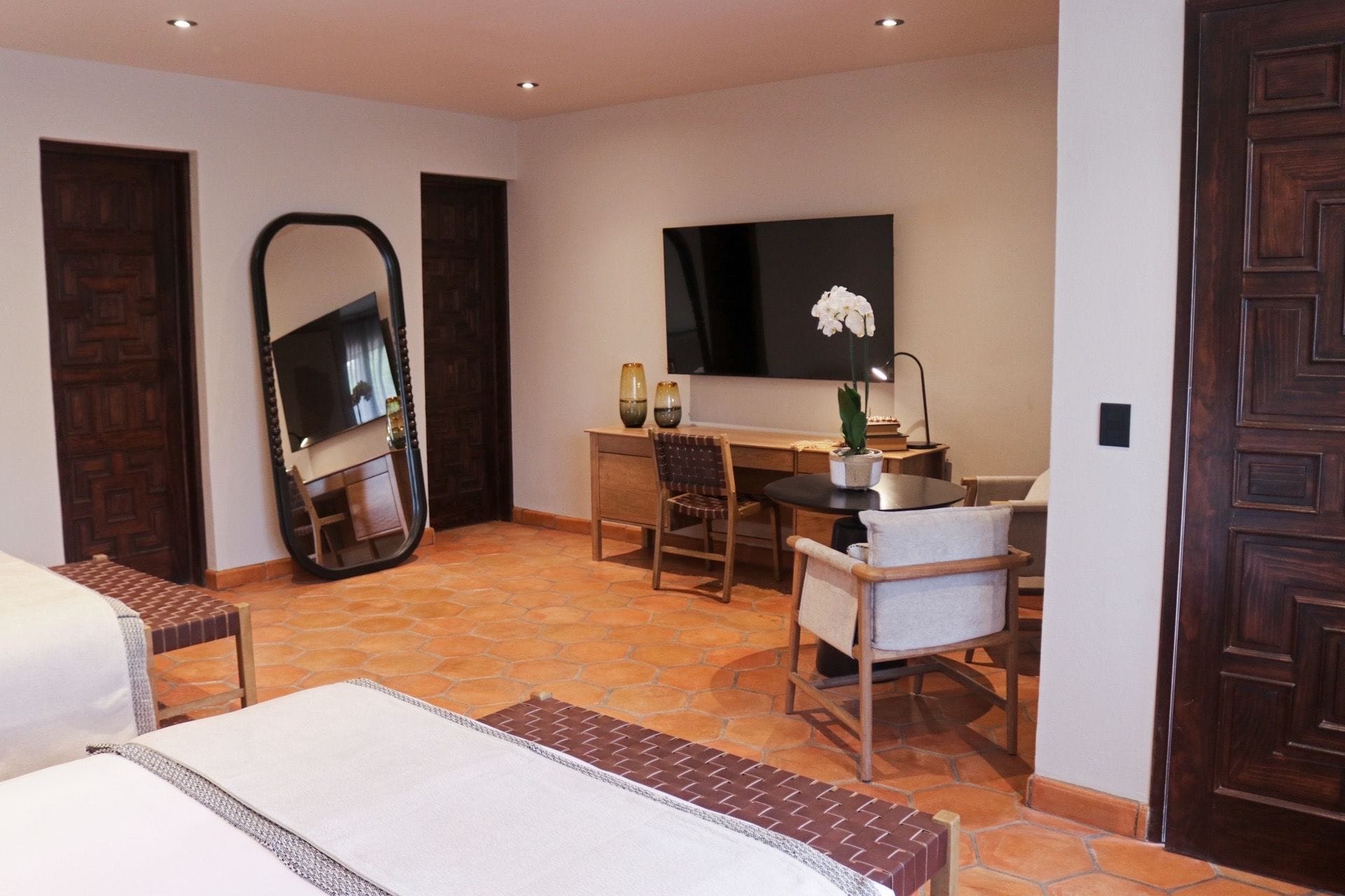 Habitación de hotel con cama, escritorio, silla, espejo grande, planta en maceta y televisor.