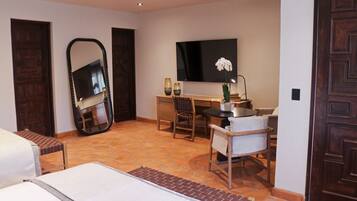 Habitación de hotel con cama, escritorio, silla, espejo grande, planta en maceta y televisor.
