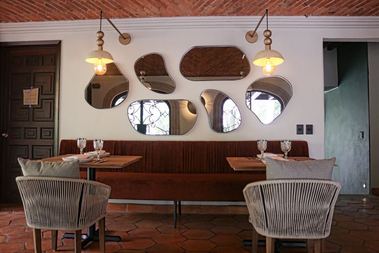 Interior de restaurante con banco de madera, sillas de mimbre y pared decorada con espejos y luces colgantes.