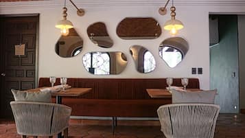 Interior de restaurante con banco de madera, sillas de mimbre y pared decorada con espejos y luces colgantes.