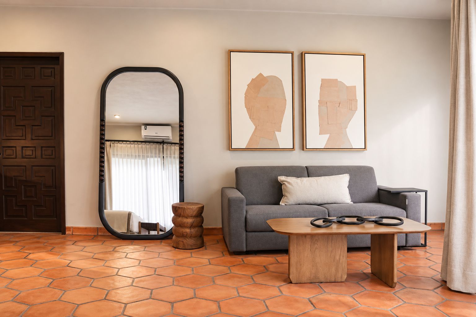 Una sala de estar con un sofá gris, una mesa redonda de madera, un espejo grande y dos obras de arte enmarcadas en la pared.