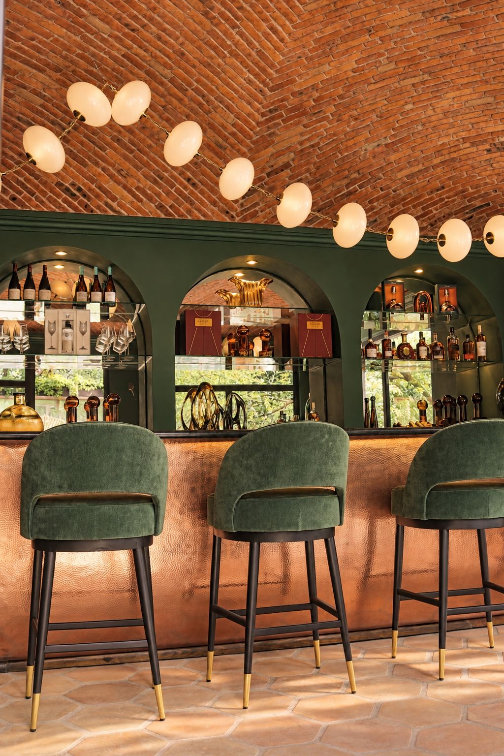 Eine Bar mit grünen, gepolsterten Barhocker, einer Ziegeldecke und einer Flaschenauslage hinter einer Glasscheibe.