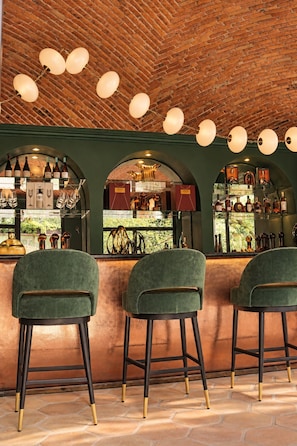 Zona de bar con taburetes tapizados en verde, techo de ladrillo y exhibición de botellas tras una mampara de cristal.