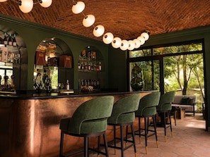 A bar area with a brick ceiling, green walls, and a large window with a view of trees. There are bar stools and a counter with bottles and glasses.