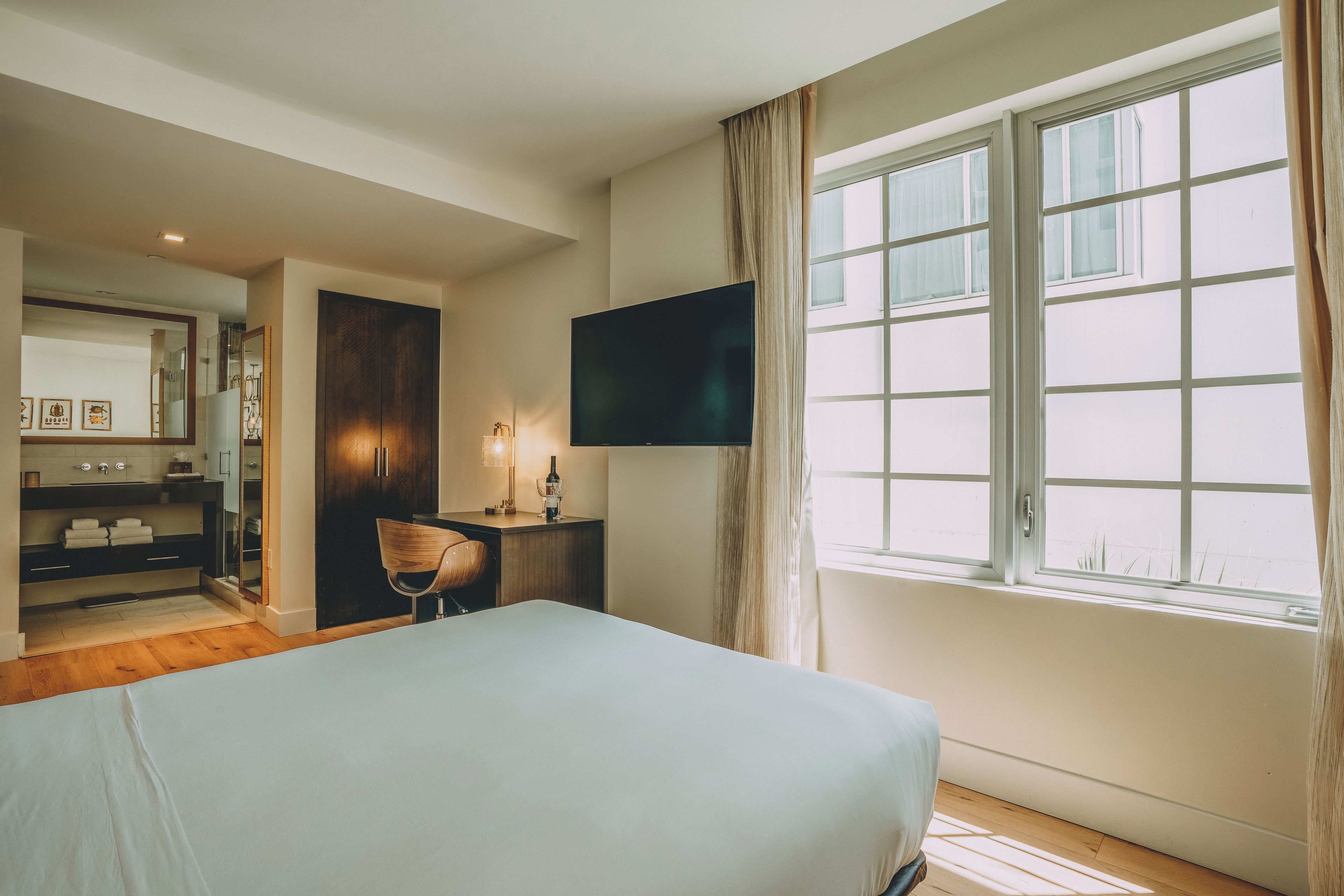 A hotel room with a large bed, a desk with a flat-screen TV, a chair, a desk lamp, and a window with curtains.