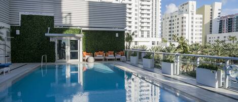 A swimming pool with a glass enclosure, outdoor seating, and a view of high-rise buildings.