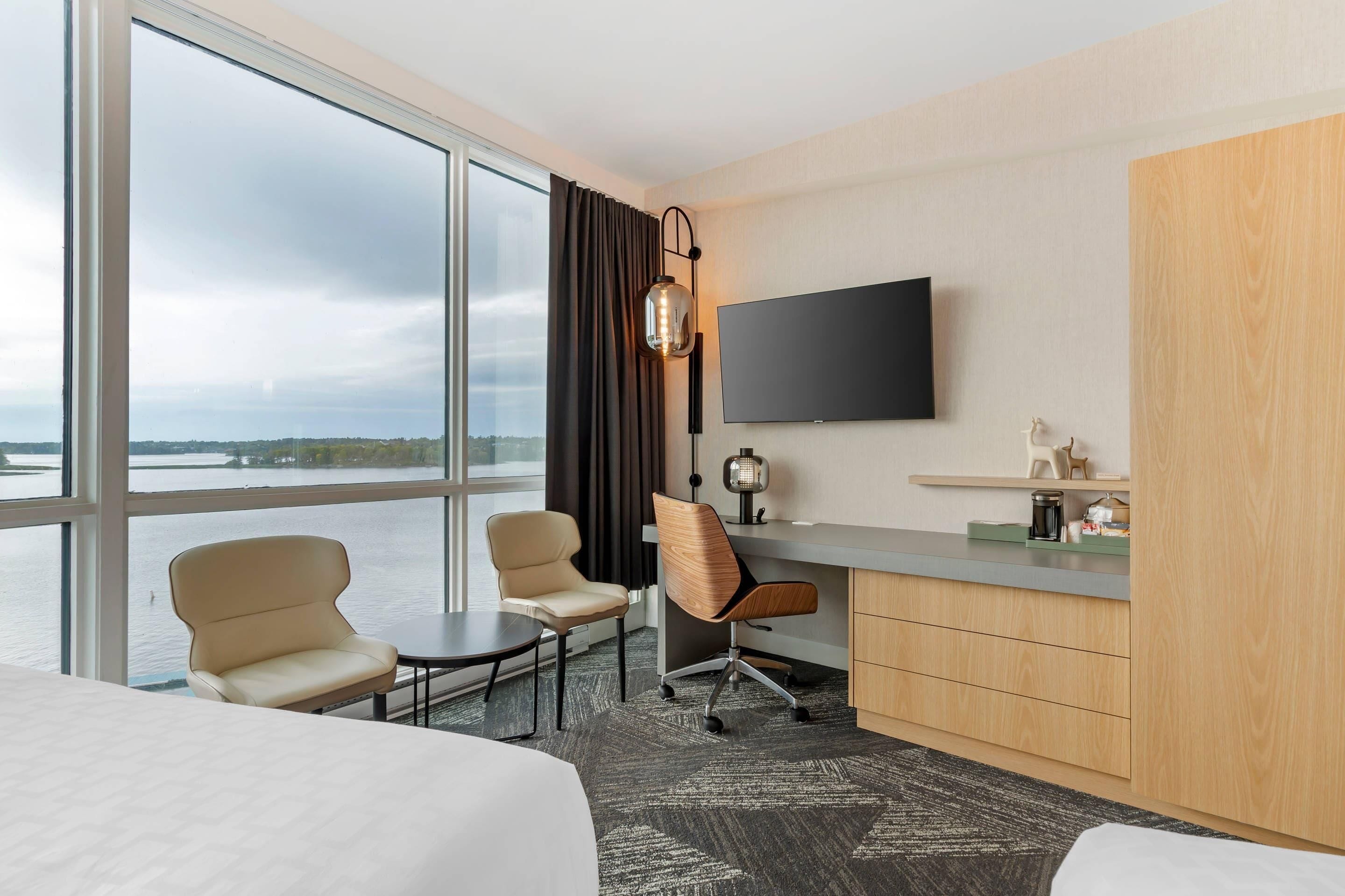 A hotel room with a large window, a desk, two chairs, a bed, and a TV.