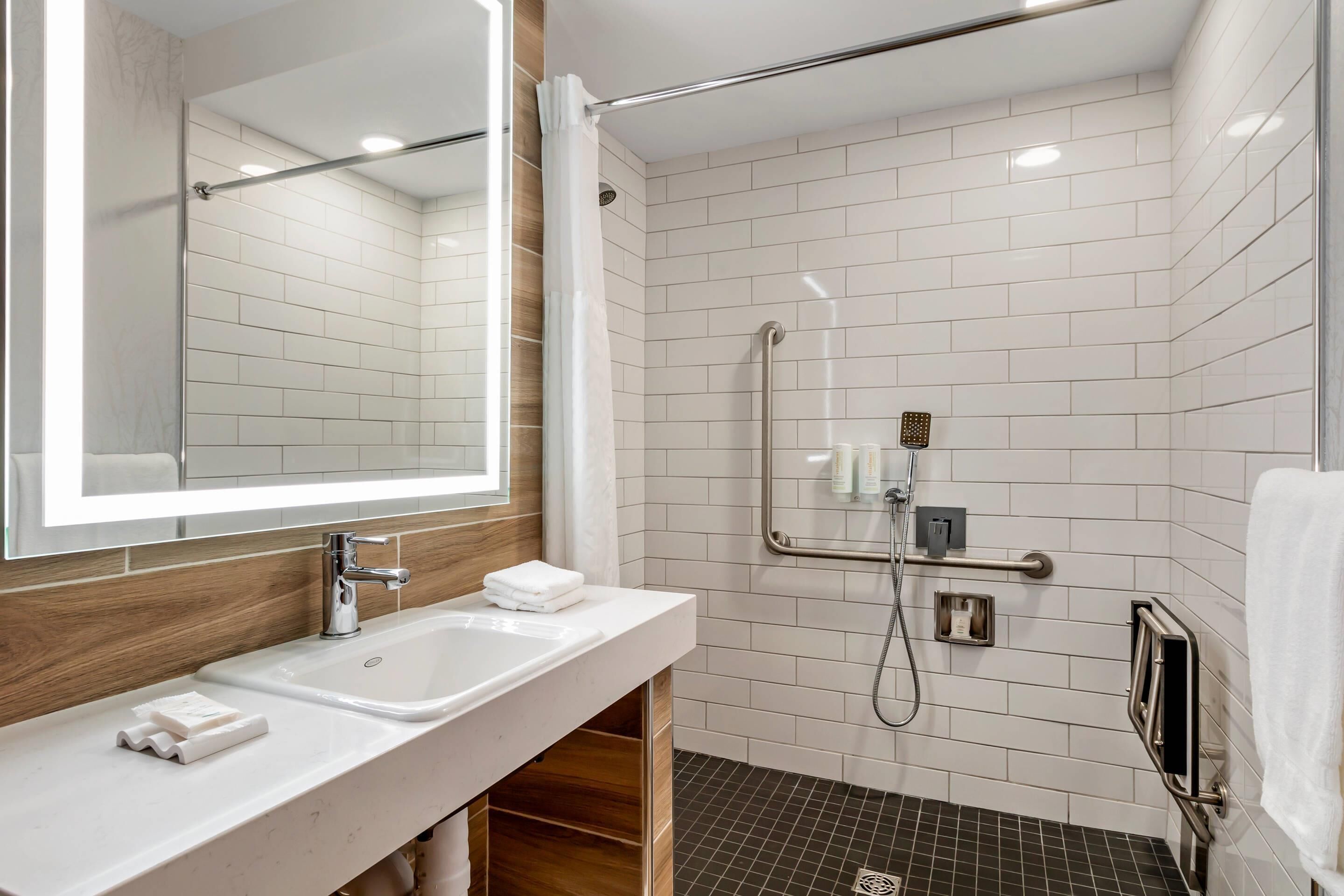 A modern bathroom with a large mirror, two sinks, a shower with grab bars, and a towel rack.