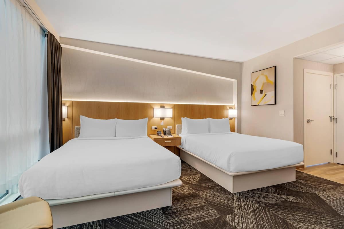 Une chambre d’hôtel avec deux lits, une table de chevet avec une lampe, une œuvre d’art accrochée au mur et une porte sur le côté droit.