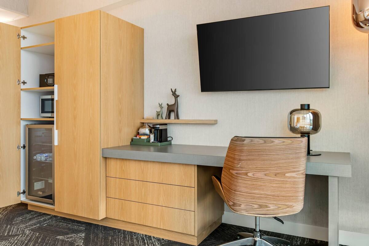 Une chambre moderne avec une armoire en bois, un bureau avec une chaise, un téléviseur fixé au mur et une lampe décorative.