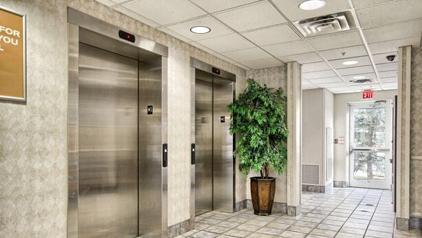A modern elevator area with metallic elevators, a potted plant, and a tiled floor. - Motel 6 London, ON - Ontario (London)