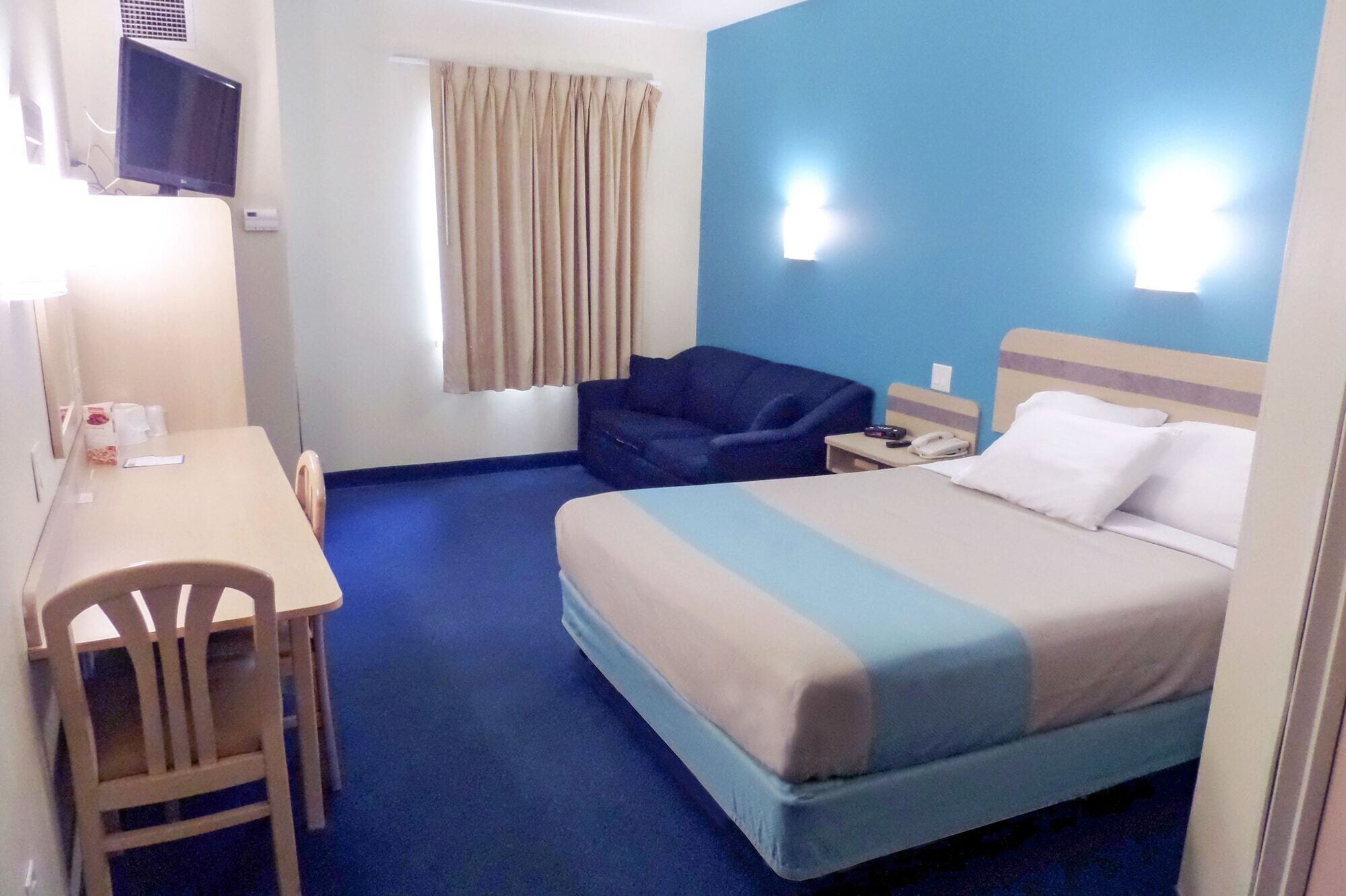 Habitación de hotel con cama, escritorio, silla, televisor y ventana con cortinas.
