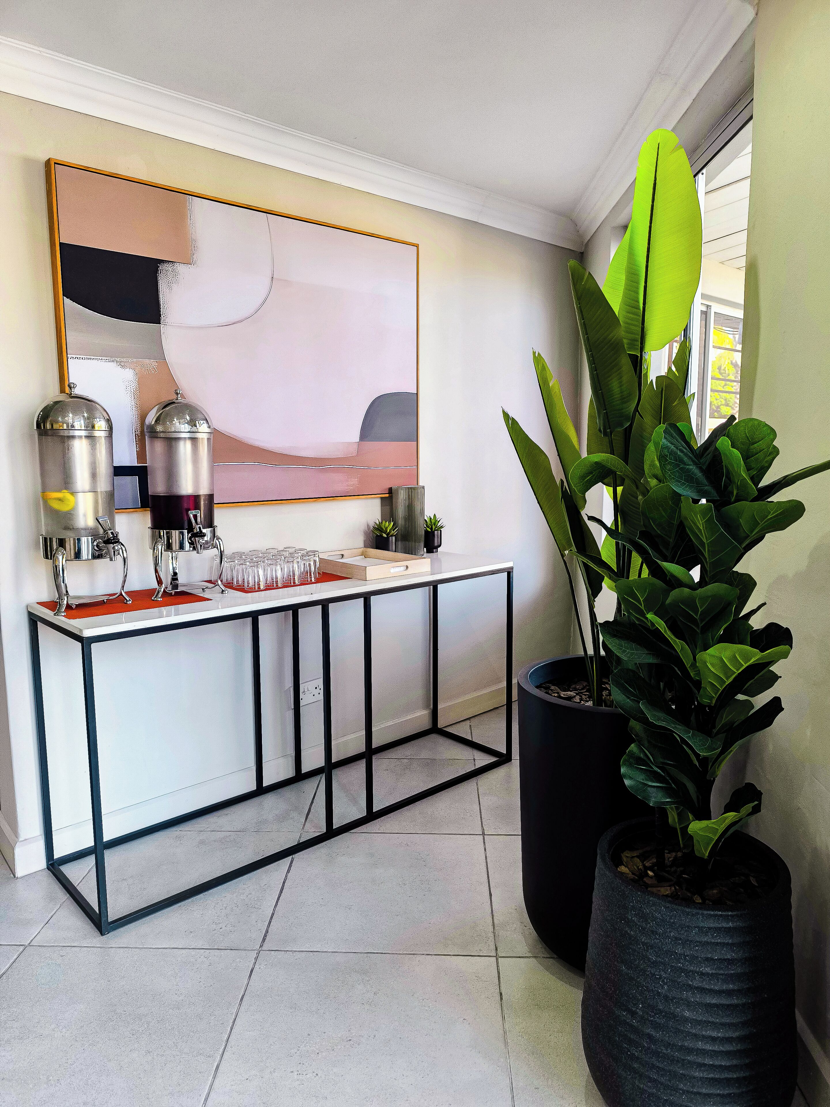 A modern kitchen with a white countertop, two stainless steel beverage dispensers, a large abstract painting, and a potted plant.