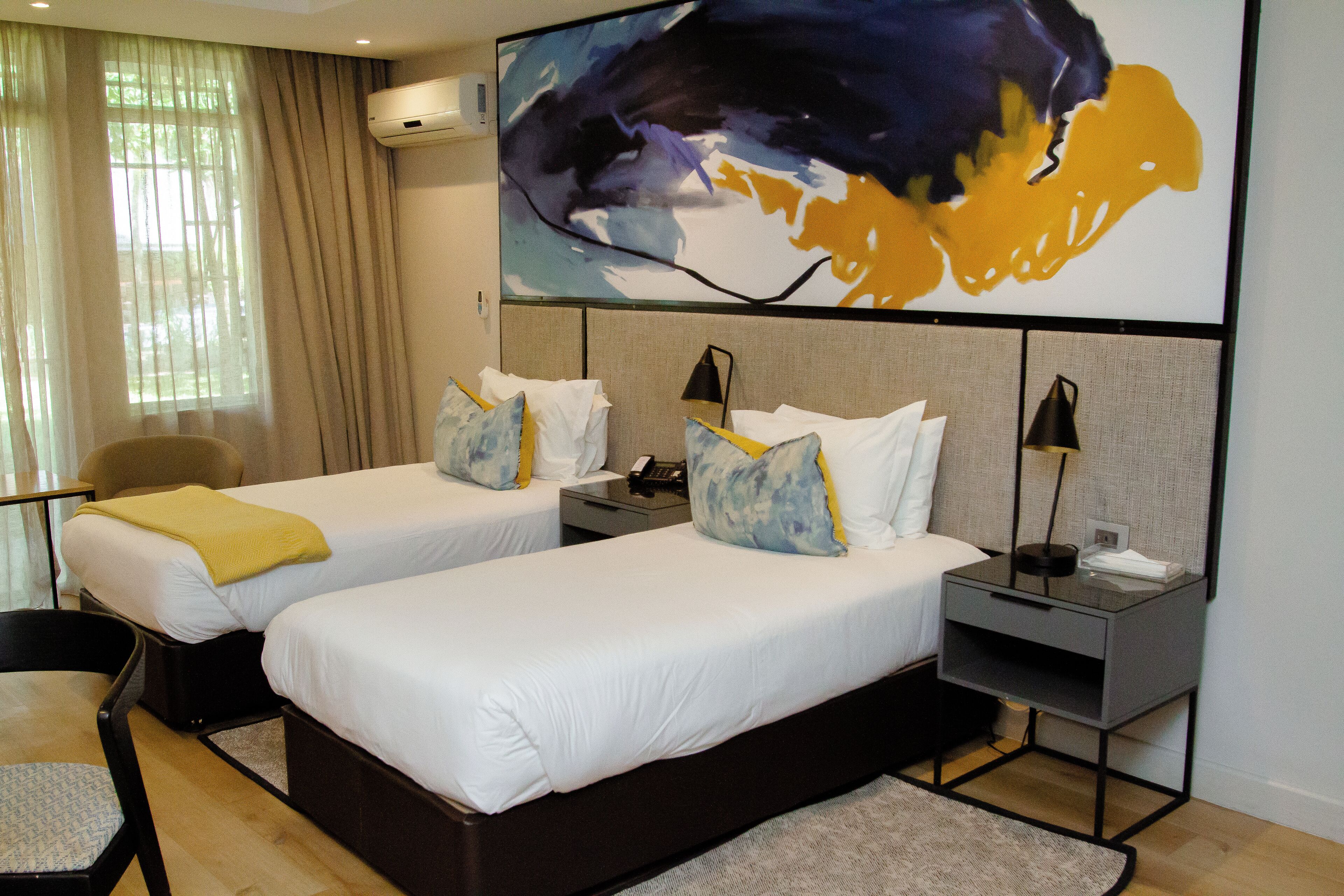 A hotel room with two single beds, a desk, a chair, and a large abstract painting on the wall.