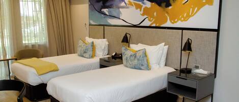 A hotel room with two single beds, a desk, a chair, and a large abstract painting on the wall.