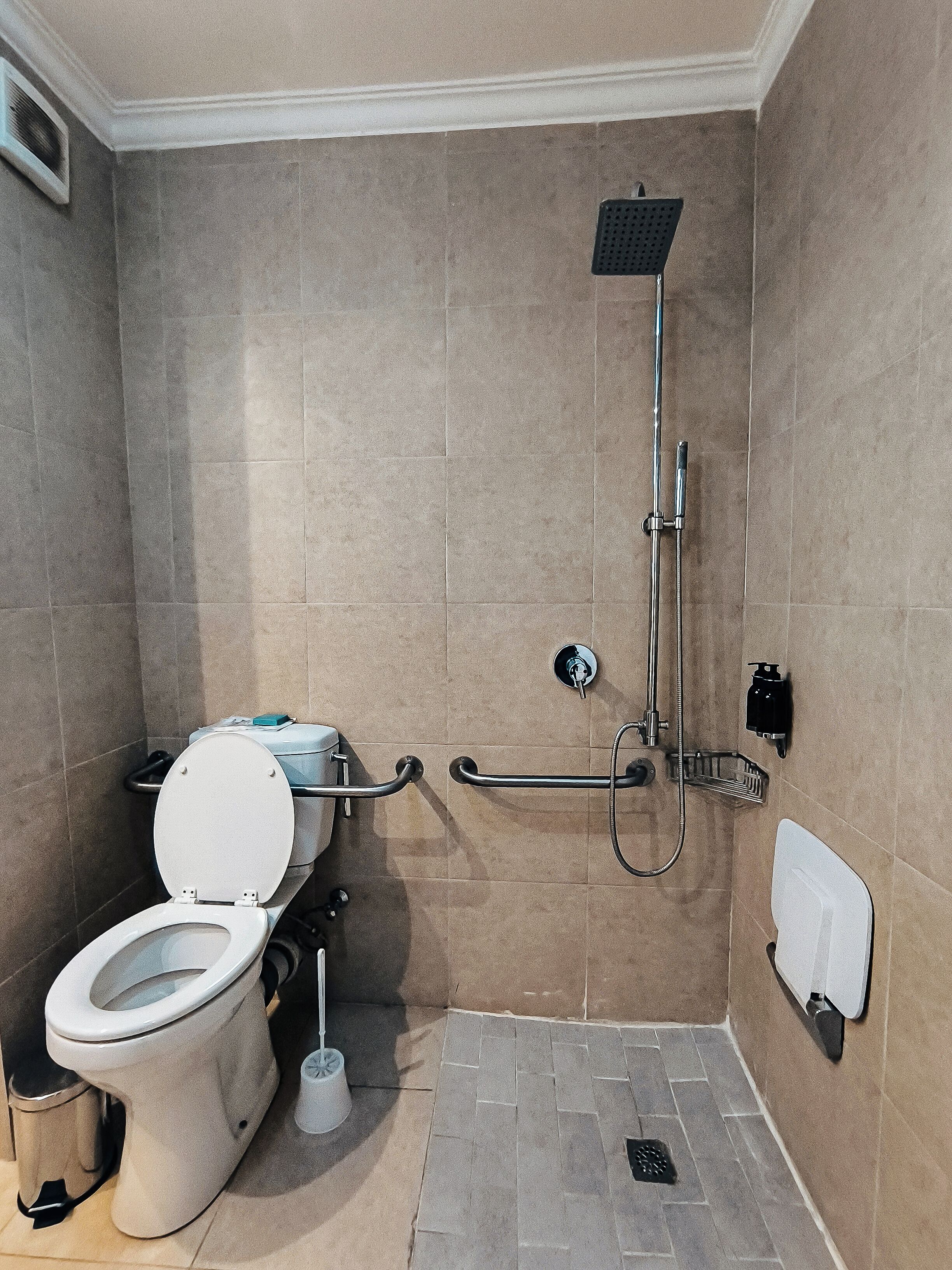 A bathroom with a toilet, shower, grab bars, and a trash can.