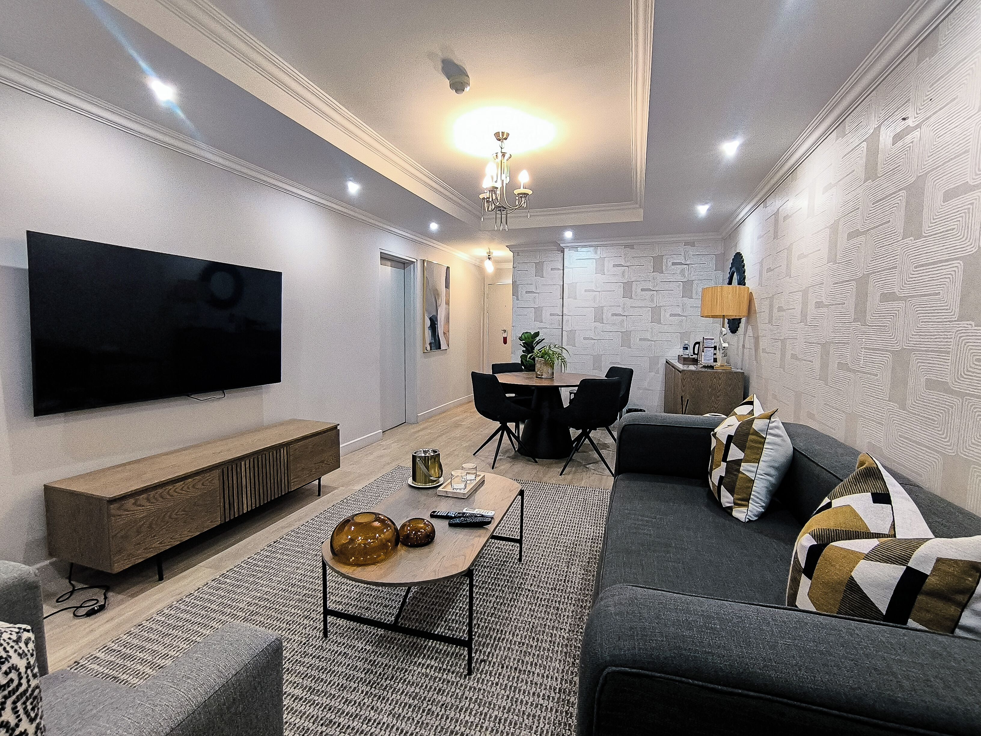 Uma sala de estar moderna com uma TV de tela plana, um espaço de jantar com mesa e cadeiras, e um sofá com almofadas estampadas.