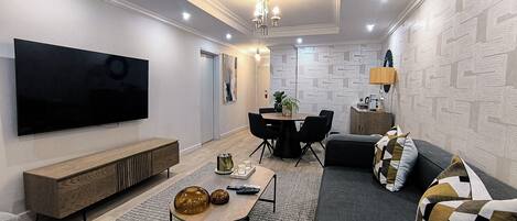 Uma sala de estar moderna com uma TV de tela plana, um espaço de jantar com mesa e cadeiras, e um sofá com almofadas estampadas.