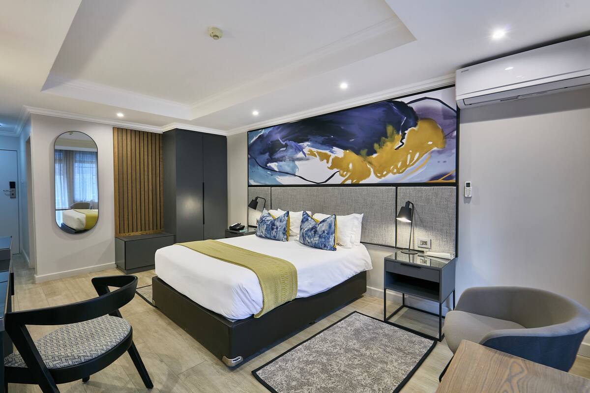 A modern hotel room with a large bed, a desk, a chair, and a large abstract painting on the wall.