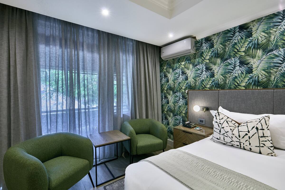 A hotel room with a bed, a green armchair, a small table, and a nightstand with a lamp.