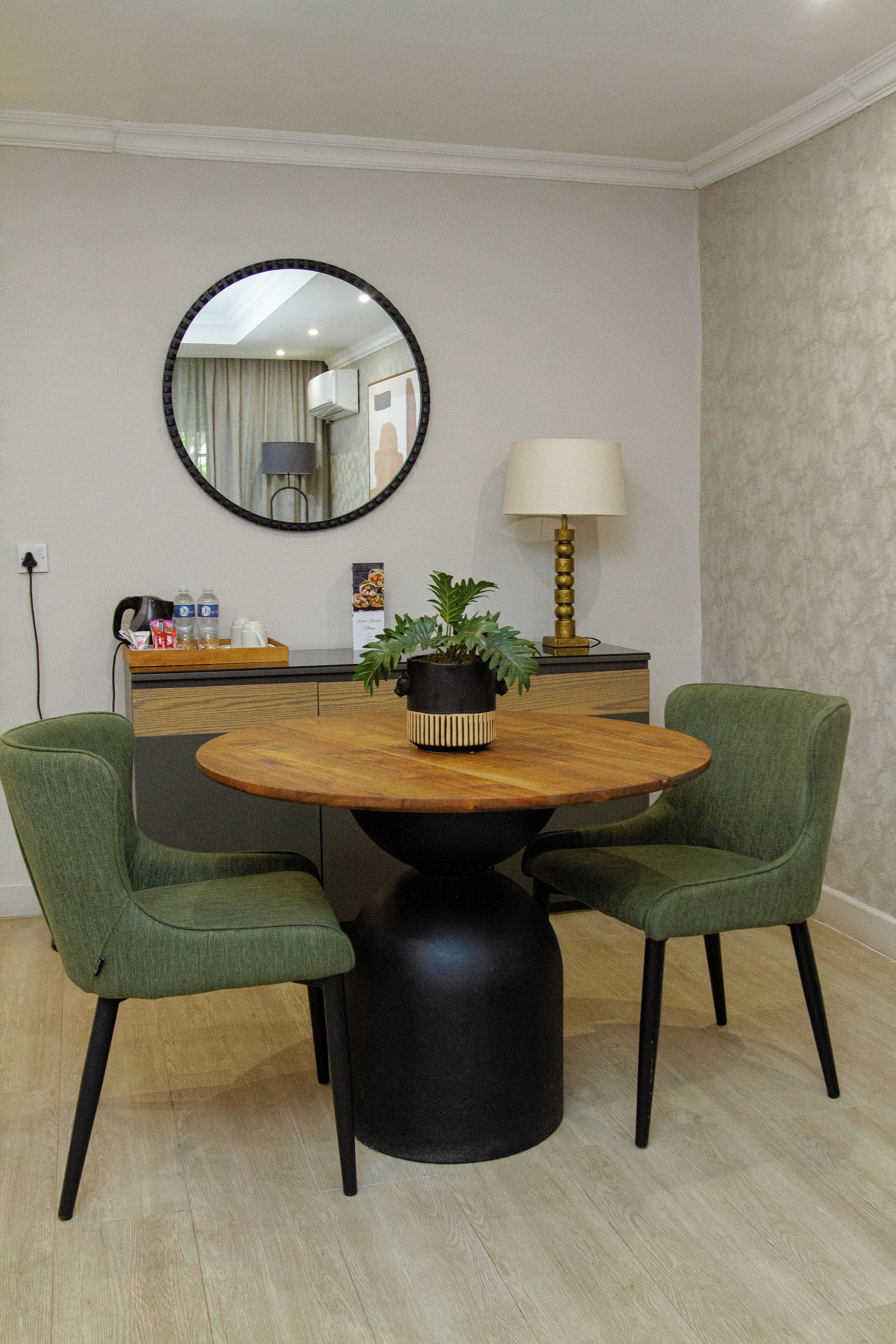 A dining area with a round wooden table, two green upholstered chairs, a black pedestal base, a potted plant, a wall-mounted mirror, a lamp, and a counter with a coffee maker and water bottles.