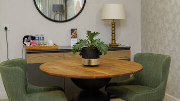 Uma área de refeições com uma mesa redonda de madeira, duas cadeiras estofadas verdes, uma base de pedestal preta, uma planta em vaso, um espelho fixado na parede, uma luminária e um balcão com cafeteira e garrafas de água.