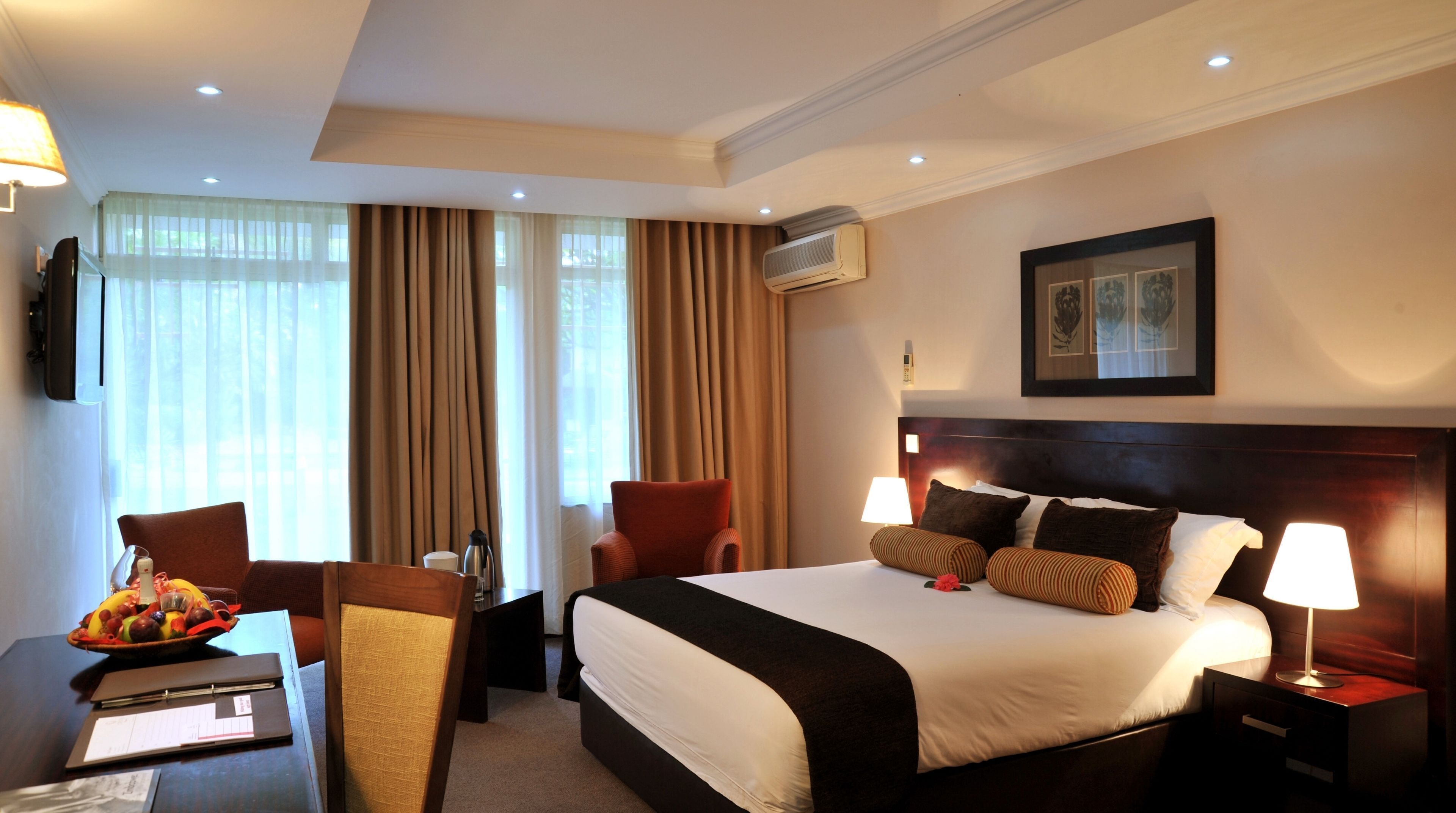 Ein Hotelzimmer mit einem Bett, zwei Stühlen, einem Schreibtisch mit einem Laptop, einem gerahmten Bild an der Wand und einer Klimaanlage.