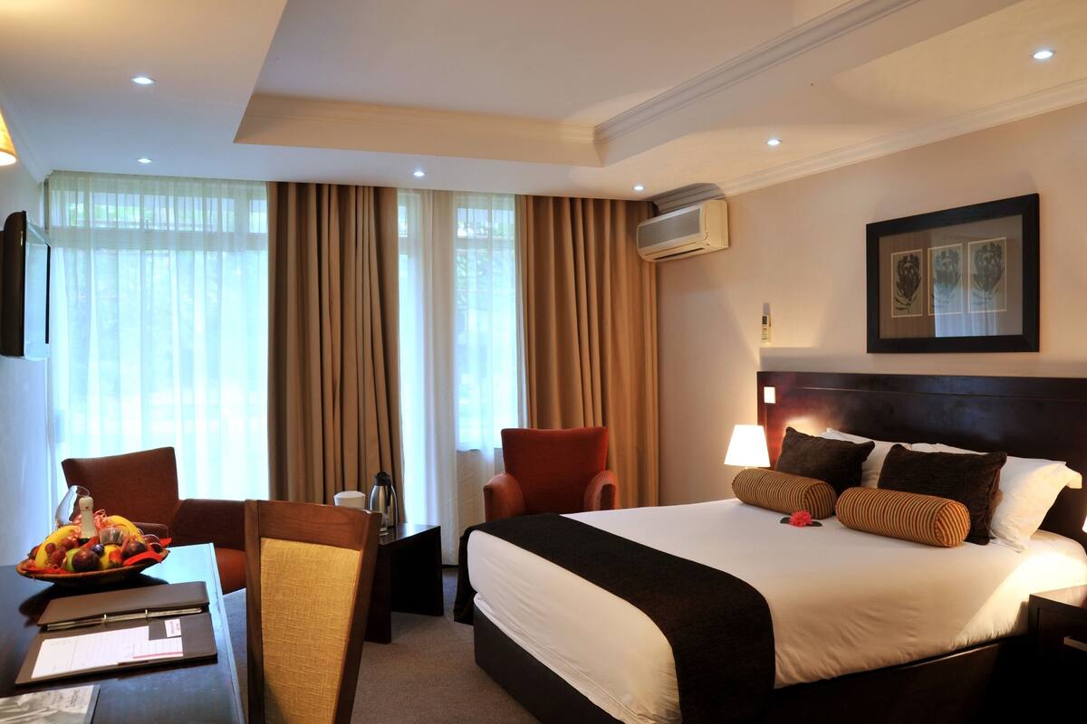 A hotel room with a bed, two chairs, a desk with a laptop, a framed picture on the wall, and an air conditioning unit.