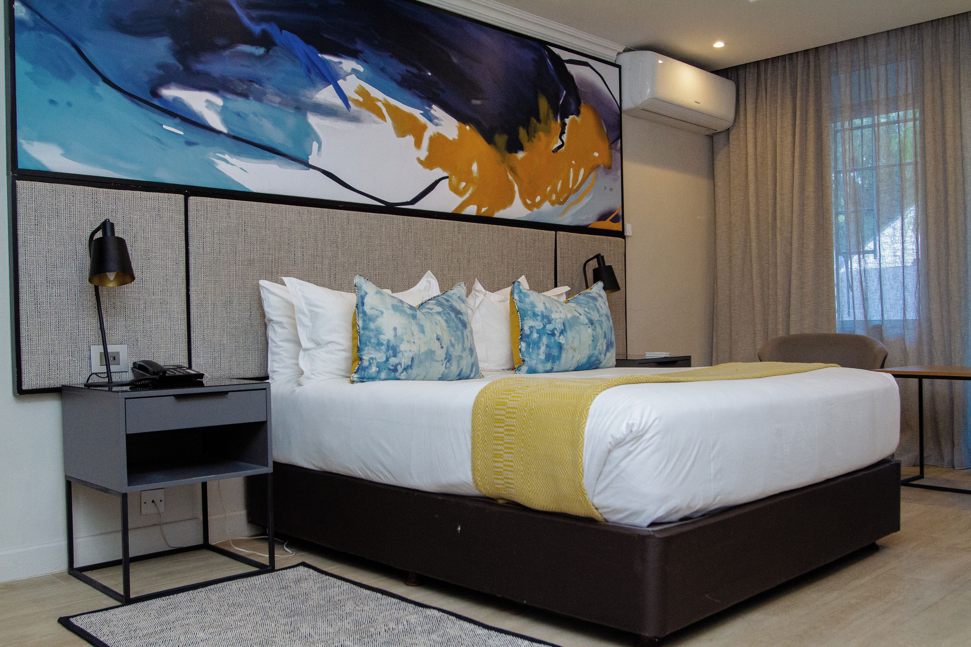 A modern hotel room with a large bed, a desk with a lamp, a large abstract painting on the wall, and a view of the outdoors through the window.