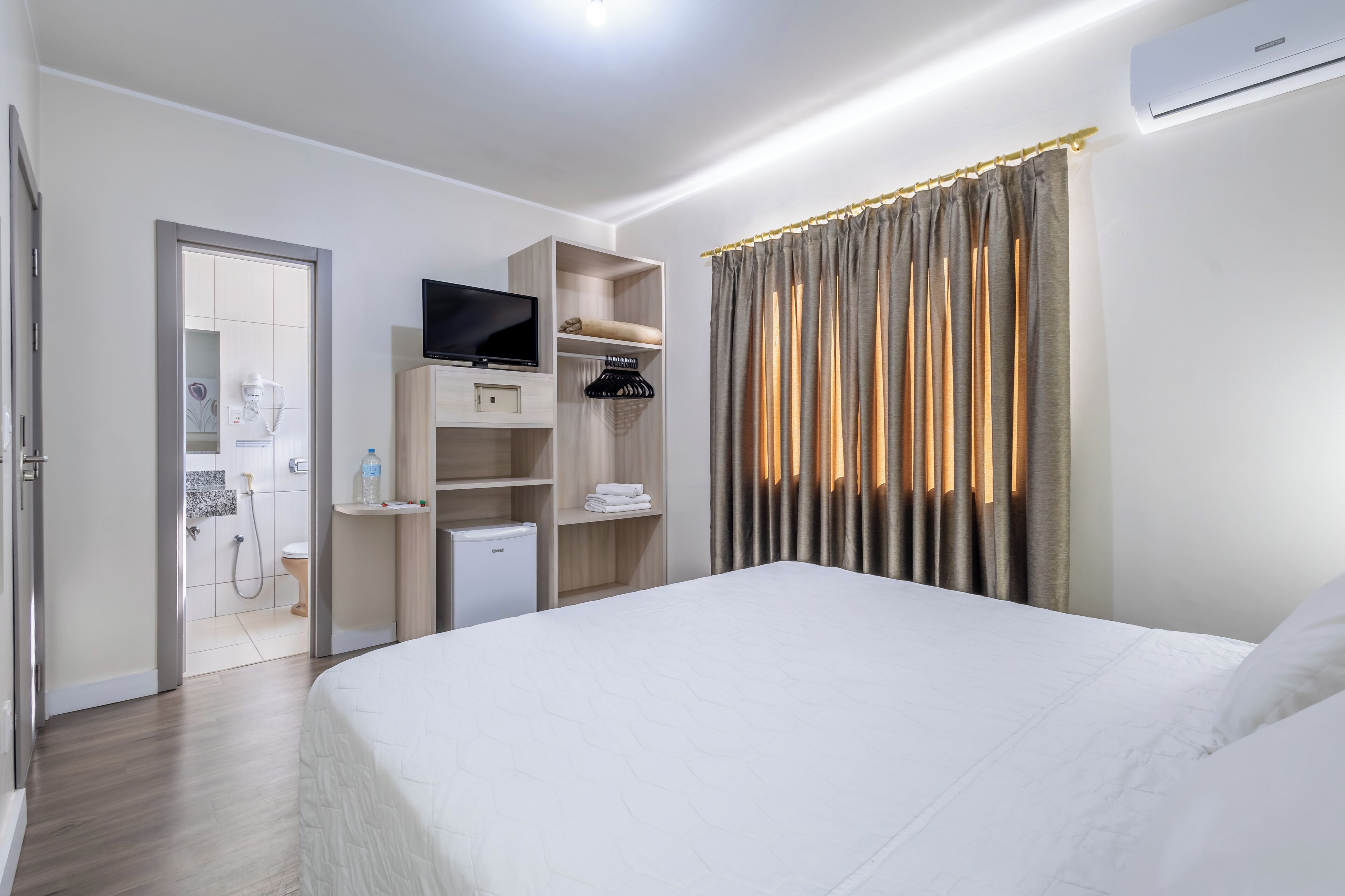 A hotel room with a bed, a TV, a mini-fridge, a shower area, and a wardrobe.