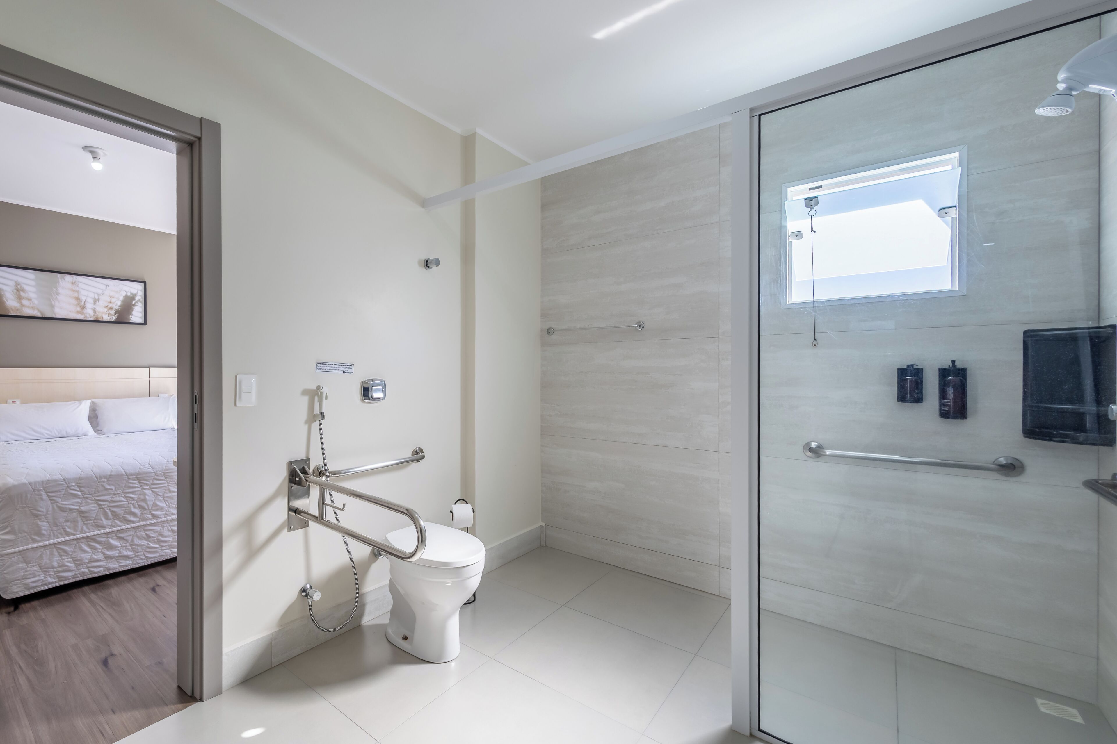 Un baño moderno con cabina de ducha de vidrio, inodoro con barra de apoyo y cama en la habitación contigua.
