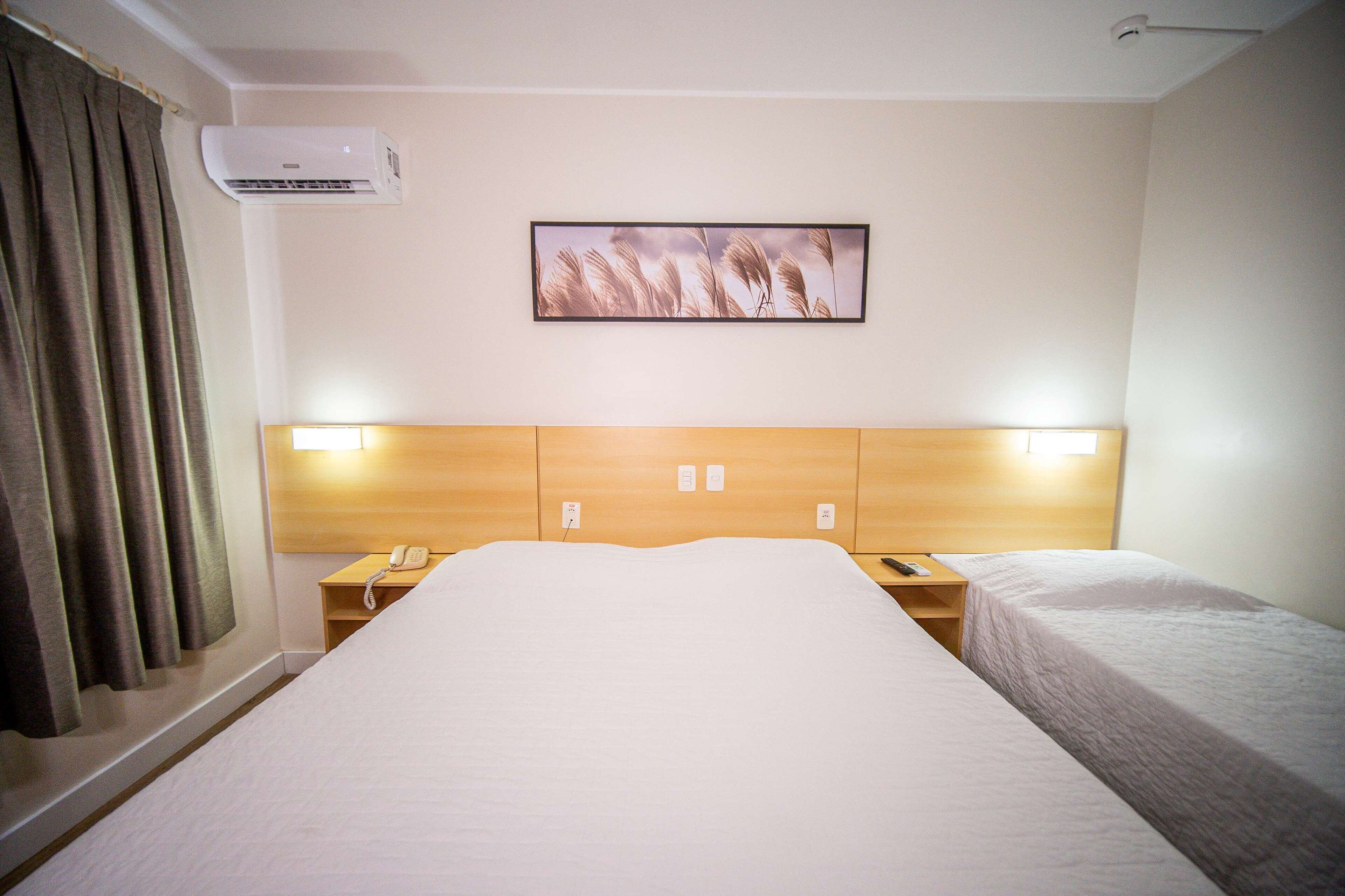 A hotel room with a large bed, a bedside table with a lamp, a phone, and a wall-mounted air conditioner.