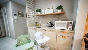 A compact kitchen with a microwave, a coffee maker, and a small shelf with plants. There are two chairs, one green and one white, and a small table.