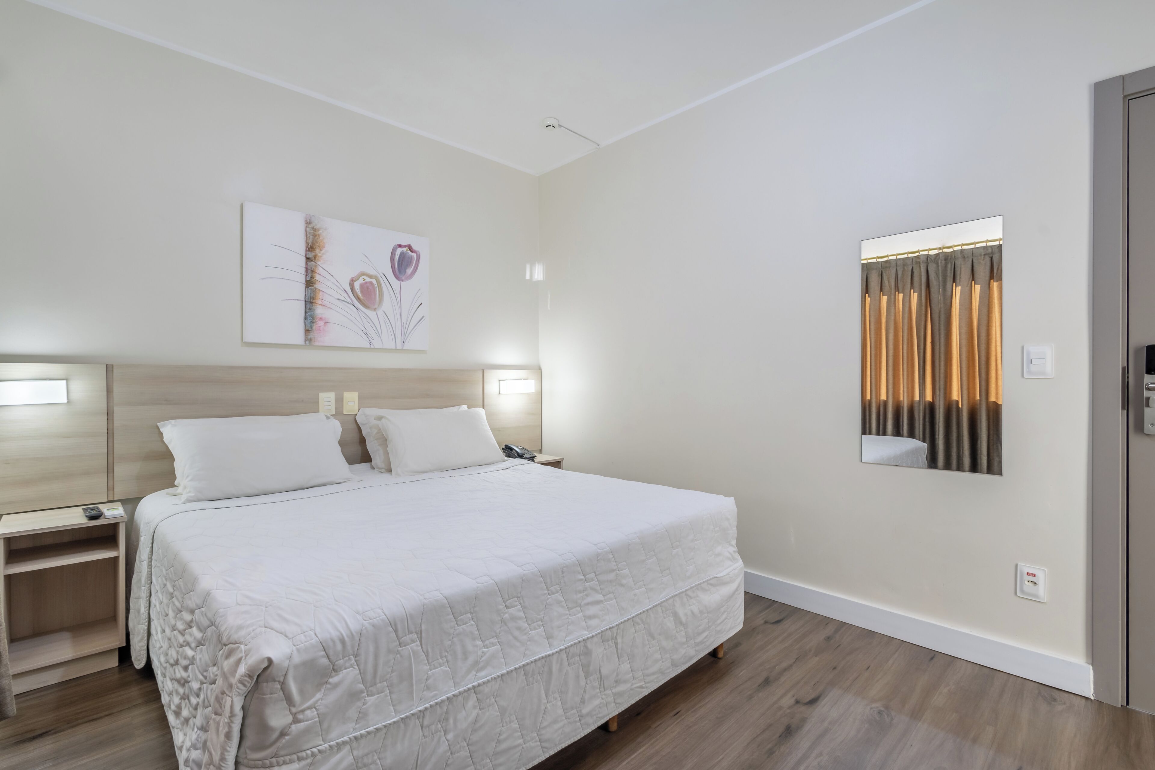 A hotel room with a large bed, bedside tables, a nightstand, a wall-mounted mirror, and a painting.